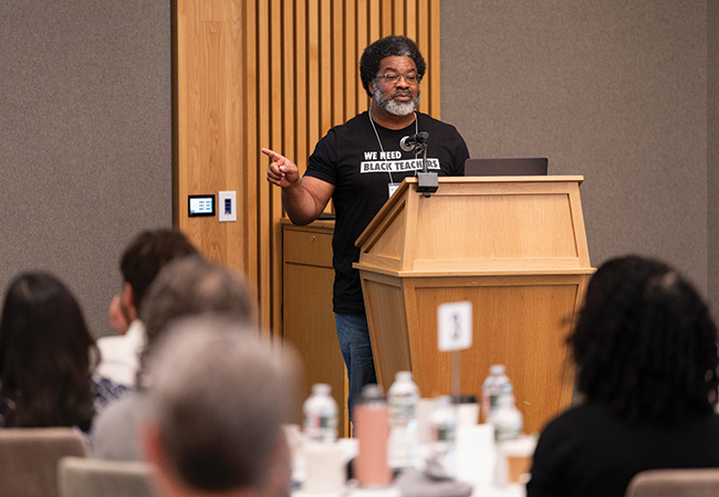 A man in a “We Need Black Teachers” t-shirt points to a screen while speaking at a podium in front of a crowd.