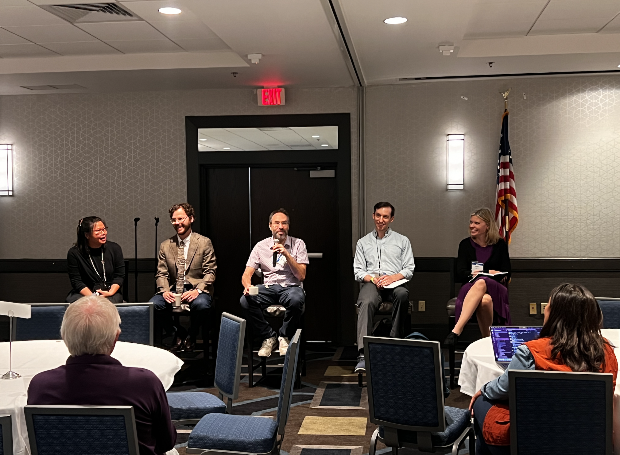 <span>Ryan Baker sits on a stool, speaking into a microphone, among other panelists in front of an audience at ICQE.</span>